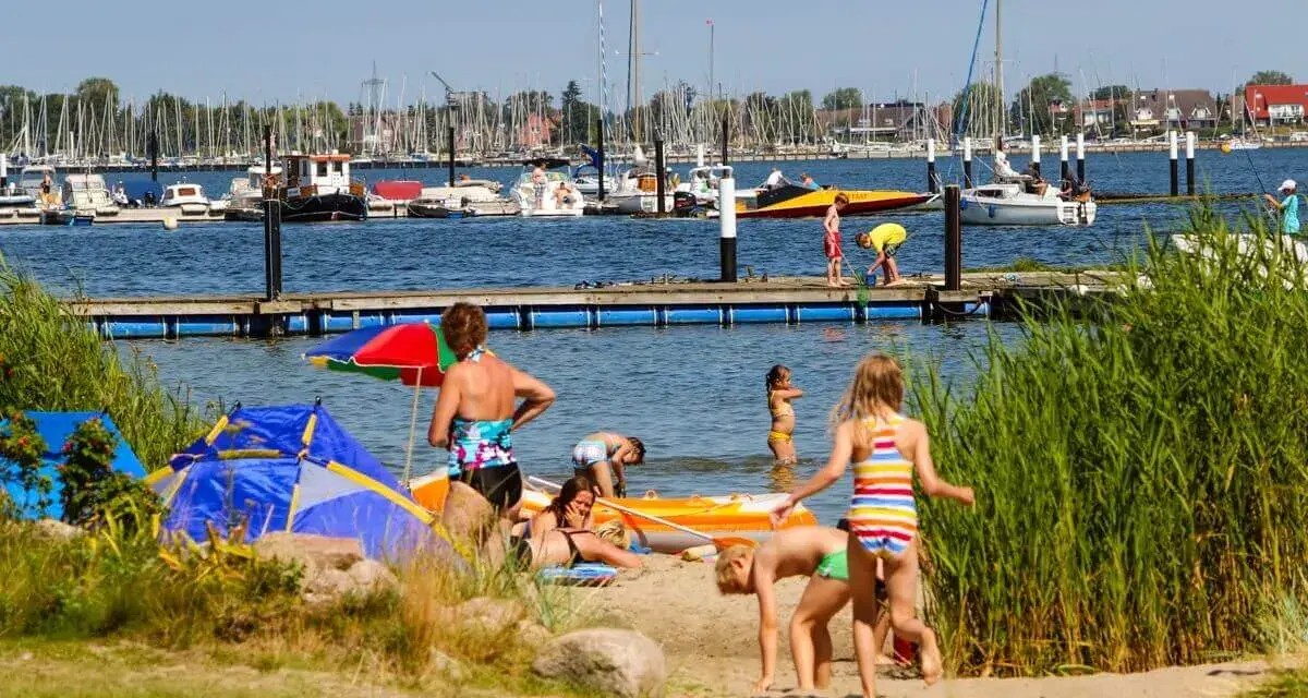 Familien genießen einen sonnigen Tag am Wasser. Kinder planschen, während Boote im Hintergrund ankern. Ein perfekter Ort für Seekamp Camping.
