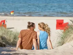 Ein Paar sitzt im Sand am Trassenheide Strand, blickt sich an. Im Hintergrund das Meer und rote Strandkörbe.