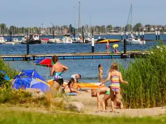 Familien genießen einen sonnigen Tag am Wasser. Kinder planschen, während Boote im Hintergrund ankern. Ein perfekter Ort für Seekamp Camping.