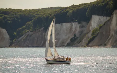Segelboot gleitet über glitzerndes Wasser vor Kreidefelsen und grünen Wäldern. Ein Tag zum Ostsee segeln.