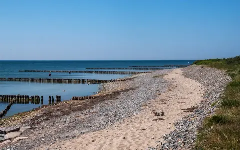Ein ruhiger Tag am Dranske Strand. Holzpfähle ragen aus dem ruhigen Meer, das sich unter einem klaren blauen Himmel erstreckt.