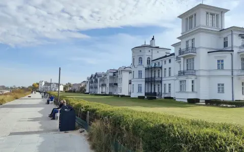 Weiße Villen säumen die Heiligendamm Promenade. Menschen spazieren und genießen die Aussicht auf das Meer.