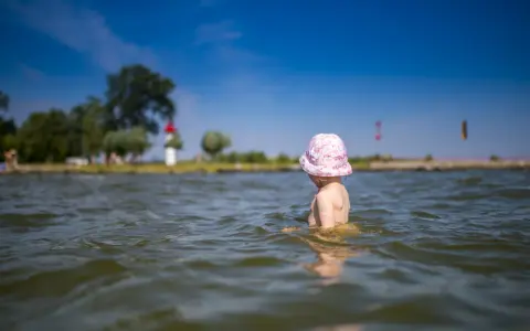 Kleines Kind mit rosa Hut planscht im Wasser am Stettiner Haff Strand. Im Hintergrund Bäume und ein Leuchtturm.