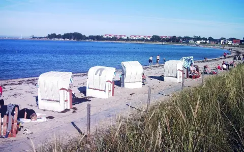 Strandkörbe am Ostseestrand, im Hintergrund der glowe hafen mit Häusern und Bäumen.