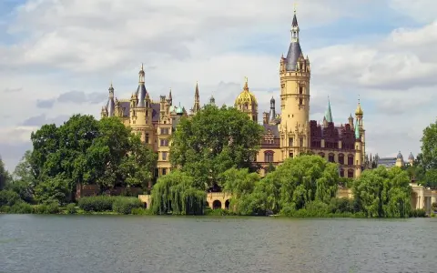 Ein märchenhaftes Schloss mit vielen Türmen und goldenen Kuppeln, umgeben von grünen Bäumen, spiegelt sich im Wasser. Ein gut gremmelin-artiger Anblick.