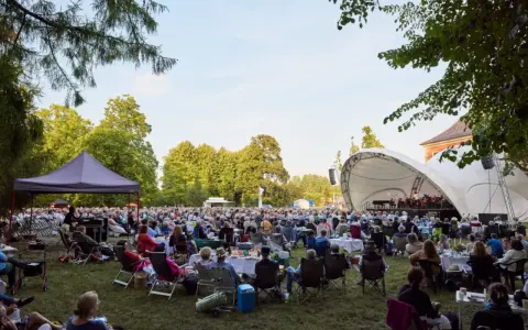 Großes Publikum bei einem Open-Air-Konzert im Rahmen eines **Festivals Mecklenburg-Vorpommern**. Eine Bühne mit Orchester, viele Sitzende auf Rasenflächen.