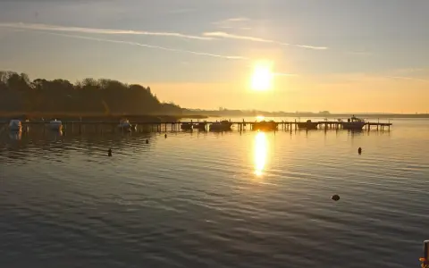 Sonnenaufgang am **Stralsund Strand**. Boote liegen am Steg, die Sonne spiegelt sich im ruhigen Wasser.