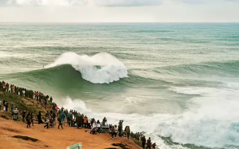 Riesige Welle bricht vor Zuschauern am mole strand. Ein Surfer reitet die Welle.
