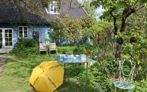 Ein Garten mit blauem Haus, gelbem Regenschirm (Kunst Offen) und Glaskunst auf einem Tisch. Pfingsten 2026 MV.