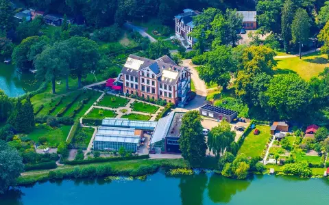 Luftaufnahme des schaugarten am tiefwarensee mit Gewächshäusern, einem Hauptgebäude und gepflegten Gärten am Ufer.