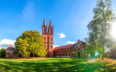 Das Kloster Dobbertin erstrahlt im Sonnenschein. Ein imposanter Kirchturm mit zwei Spitzen ragt empor, umgeben von grünen Rasenflächen und alten Bäumen.