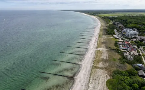 Luftaufnahme des Ahrenshoop Strandes mit Wellenbrechern, Sand und Bäumen. Ein Segelboot ist auf dem Meer zu sehen.