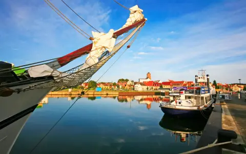 Ein Segelschiff im Vordergrund, im Hintergrund die Stadt Wolgast mit roten Dächern und einem Turm. Ein kleines Boot mit dem Namen "Wolgast" liegt am Kai.