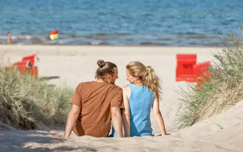 Ein Paar sitzt im Sand am Trassenheide Strand, blickt sich an. Im Hintergrund das Meer und rote Strandkörbe.
