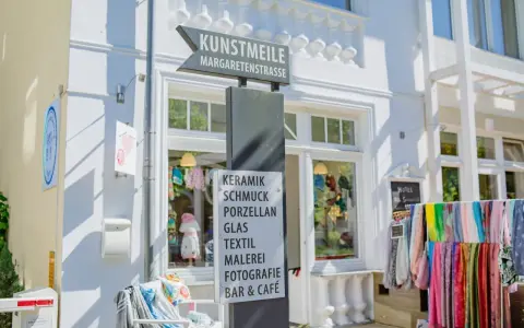 Schild an der Kunstmeile Binz: Keramik, Schmuck, Porzellan, Glas, Textil, Malerei, Fotografie, Bar & Café. Bunte Schals hängen davor.