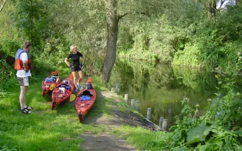 Am Ufer der Kanustation Mirow stehen drei rote Kajaks bereit. Ein Mann mit Schwimmweste und eine Frau in Sportkleidung warten auf ihre Paddeltour.