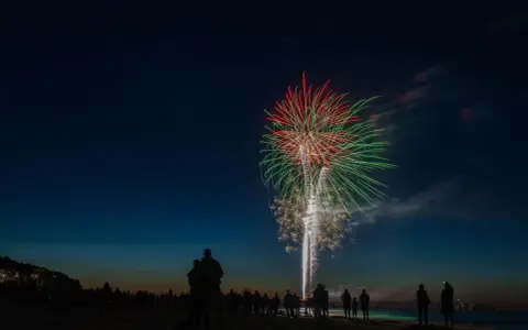 Silhouetten von Menschen am Strand beobachten ein beeindruckendes Feuerwerk über dem Meer. Ein leuchtendes Spektakel in Rot und Grün erhellt den Nachthimmel. Feuerwerk Usedom.