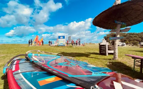 Windsurfausrüstung liegt am ummanz strand. Im Hintergrund bunte Segel und Menschen, die sich auf das Wasser vorbereiten.
