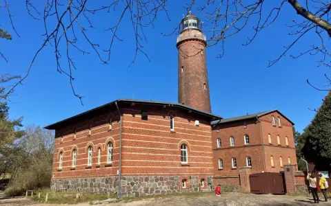 Der Leuchtturm Prerow ragt imposant in den blauen Himmel. Ziegelbauten mit Rundbogenfenstern säumen seinen Fuß.