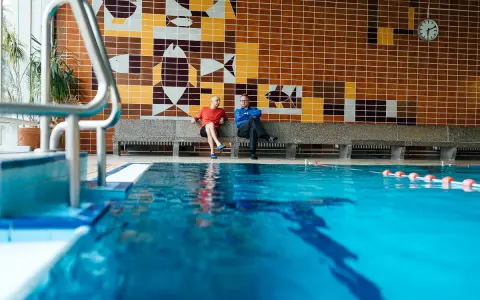 Zwei Männer sitzen in der Schwimmhalle Anklam auf einer Bank vor einer bunten Kachelwand mit Fischmotiven.