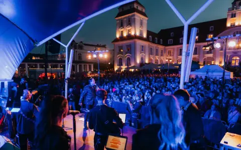 Konzert mit Sänger und Band auf der Bühne, beleuchtet von blauen Lichtern. Im Hintergrund die beleuchtete Architektur und eine Menschenmenge bei Veranstaltungen am Binz Kurplatz.