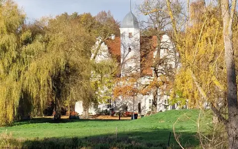 Herbstliche Farben umgeben das Wasserschloss Quilow. Ein Teich spiegelt die Bäume und das weiße Gebäude mit seinem markanten Turm.