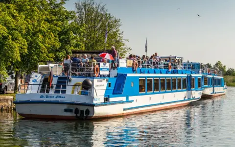 Fahrgastschifffahrt Wichmann: Zwei blau-weiße Schiffe mit Passagieren auf dem Wasser, umgeben von grünen Bäumen und klarem Himmel.