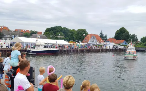 Menschen genießen das bunte Treiben beim Rock am Hafen. Boote liegen im Wasser, ein Rettungsboot fährt vorbei.