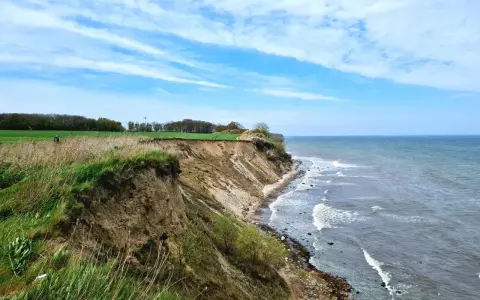 Steilküste mit grasbewachsenem Abhang, der zum steinbeck Strand und dem Meer abfällt. Wolken ziehen über den blauen Himmel.
