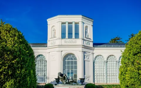 Die Orangerie Putbus, ein strahlend weißes Gebäude mit Skulpturen und großen Fenstern, wird von üppigem Grün eingerahmt.