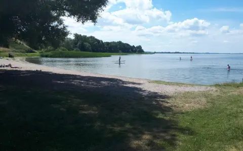 Sonniger Tag am Kummerower See. Menschen entspannen am Strand, andere paddeln auf SUPs. Herrlicher See- und Waldblick.