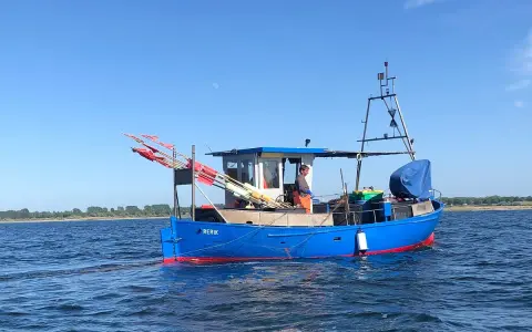 Blaues Fischerboot "RERIK" mit roten Markierungen auf dem Wasser. Ein Mann steht an Deck, rote Fahnen ragen empor.