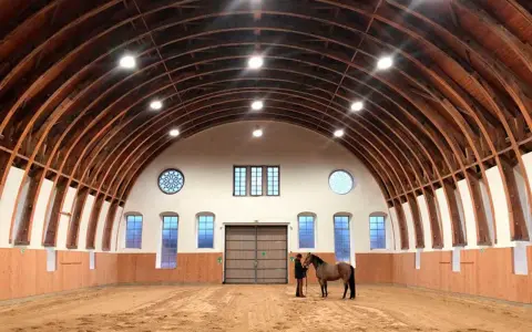 Reithalle mit Holzbalken-Decke und Sandboden. Ein Reiter steht neben seinem Pferd. Ein Ort, der an den farmer hotel Basedow erinnert.
