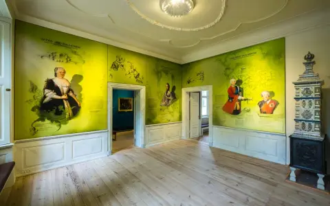 Ein Raum im Schloss Mirow mit Wandmalereien, die historische Persönlichkeiten und Karten zeigen. Ein Kachelofen steht rechts.