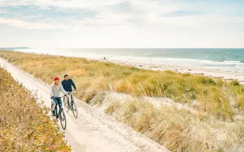 Ein Paar genießt seine baltischen Abenteuer auf Fahrrädern entlang eines sandigen Weges am Meer, umgeben von Dünen und Gräsern.