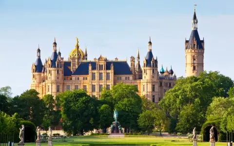 Das Schweriner Schloss, ein Märchenschloss mit goldenen Kuppeln und Türmen, thront majestätisch über einem grünen Park mit Statuen und einem Teich.