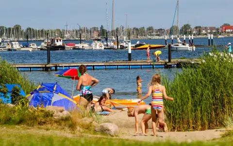 Familien genießen einen sonnigen Tag am Wasser. Kinder planschen, während Boote im Hintergrund ankern. Ein perfekter Ort für Seekamp Camping.