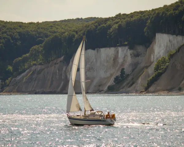 Segelboot gleitet über glitzerndes Wasser vor Kreidefelsen und grünen Wäldern. Ein Tag zum Ostsee segeln.