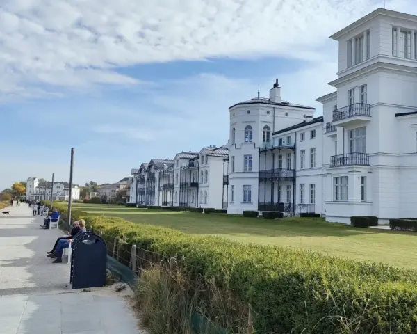 Weiße Villen säumen die Heiligendamm Promenade. Menschen spazieren und genießen die Aussicht auf das Meer.