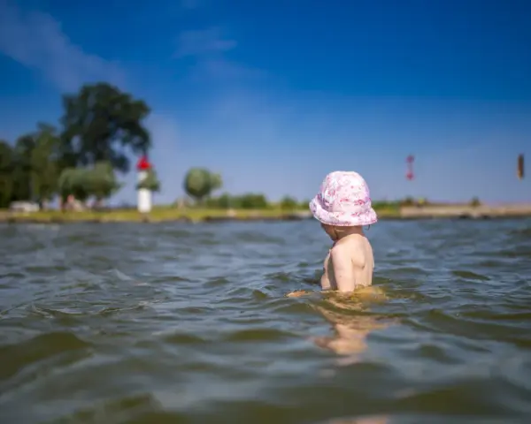 Kleines Kind mit rosa Hut planscht im Wasser am Stettiner Haff Strand. Im Hintergrund Bäume und ein Leuchtturm.