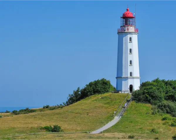 Der Leuchtturm Dornbusch ragt auf einer grünen Anhöhe empor, mit dem blauen Meer im Hintergrund. Eine Treppe führt zum Eingang.
