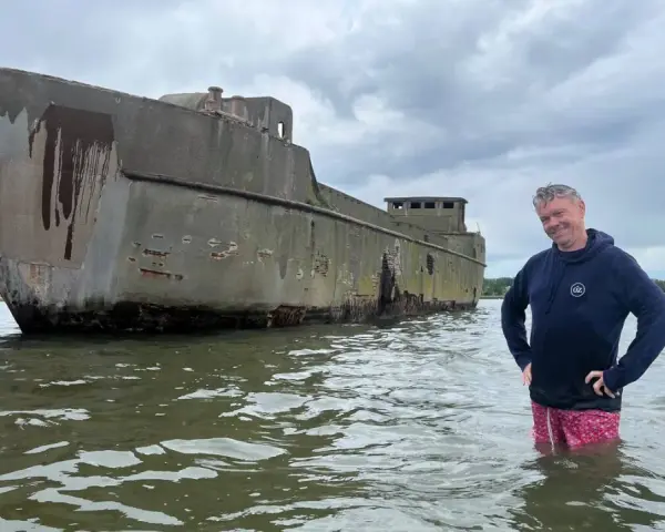Mann steht im Wasser neben einem verwitterten Betonschiff in Wismar.