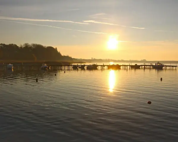 Sonnenaufgang am **Stralsund Strand**. Boote liegen am Steg, die Sonne spiegelt sich im ruhigen Wasser.