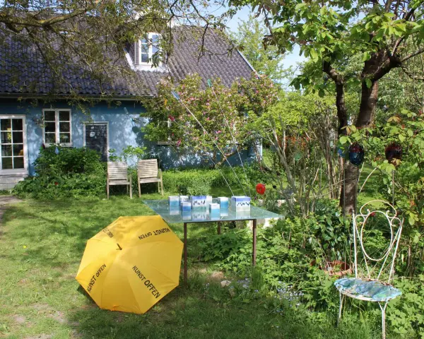Ein Garten mit blauem Haus, gelbem Regenschirm (Kunst Offen) und Glaskunst auf einem Tisch. Pfingsten 2026 MV.