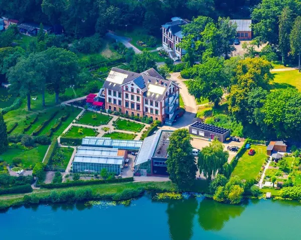 Luftaufnahme des schaugarten am tiefwarensee mit Gewächshäusern, einem Hauptgebäude und gepflegten Gärten am Ufer.