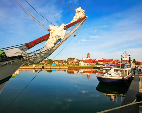 Ein Segelschiff im Vordergrund, im Hintergrund die Stadt Wolgast mit roten Dächern und einem Turm. Ein kleines Boot mit dem Namen "Wolgast" liegt am Kai.