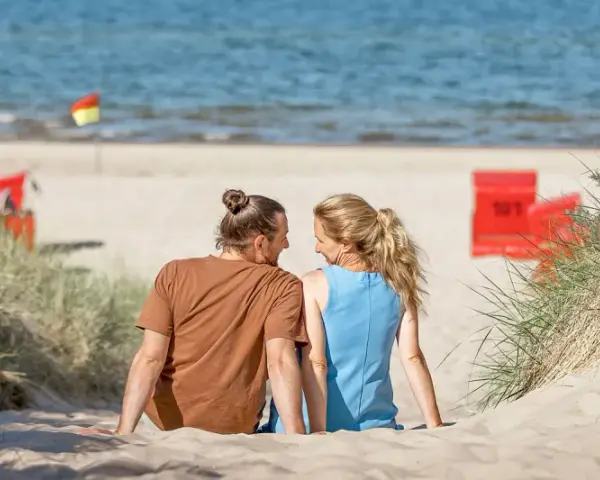 Ein Paar sitzt im Sand am Trassenheide Strand, blickt sich an. Im Hintergrund das Meer und rote Strandkörbe.