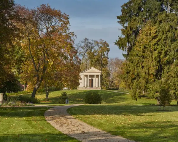 Herbstlicher Spaziergang im Schlossgarten Neustrelitz. Ein Weg führt zu einem antiken Mausoleum, umgeben von bunten Bäumen.