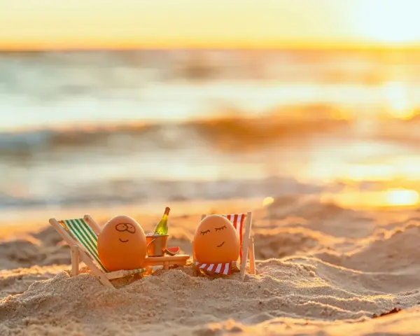 Zwei bemalte Eier entspannen sich in Liegestühlen am Strand bei Sonnenuntergang. Eine Flasche Sekt steht bereit. Eine süße Idee für Veranstaltungen an Ostern.