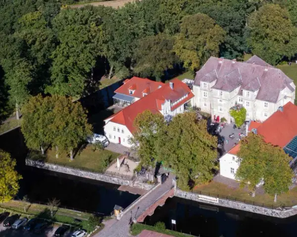 Luftaufnahme des Wasserschlosses Mellenthin mit Wassergraben, Brücke und umliegendem Park. Der Eintritt ist frei zugänglich.